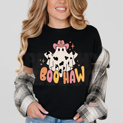 A woman wearing a black graphic t-shirt with a cartoon ghost cowboy design and the text 'BOO HAW'. She has a gray and white plaid flannel shirt draped off her shoulders and is wearing blue jeans against a light gray background.