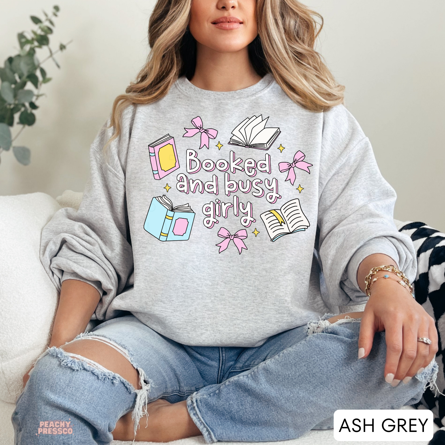 Woman wearing an ash grey crewneck sweatshirt featuring a colorful "Booked and Busy Girly" graphic with pink bows, pastel books, open books, and sparkles. Cozy oversized fit styled with distressed light wash jeans.