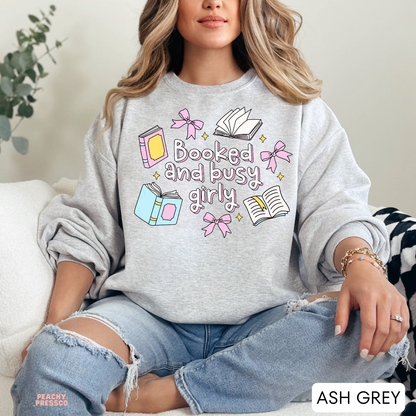 Woman wearing an ash grey crewneck sweatshirt featuring a colorful "Booked and Busy Girly" graphic with pink bows, pastel books, open books, and sparkles. Cozy oversized fit styled with distressed light wash jeans.