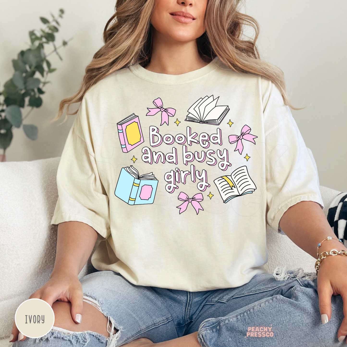 Woman wearing an ivory Comfort Colors t-shirt with a whimsical "Booked and Busy Girly" graphic featuring pastel books, pink bows, open books, and sparkles. Casual oversized bookish aesthetic.