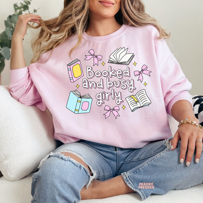 Woman wearing a light pink crewneck sweatshirt featuring a cute "Booked and Busy Girly" design with pastel books, pink bows, open books, and sparkles. Soft oversized fit styled with distressed denim.