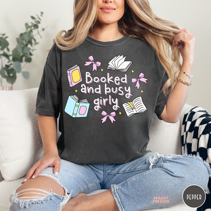 Woman wearing a pepper Comfort Colors t-shirt with a pastel "Booked and Busy Girly" book lover graphic featuring pink bows, books, open pages, and stars. Relaxed oversized fit with light wash ripped jeans.