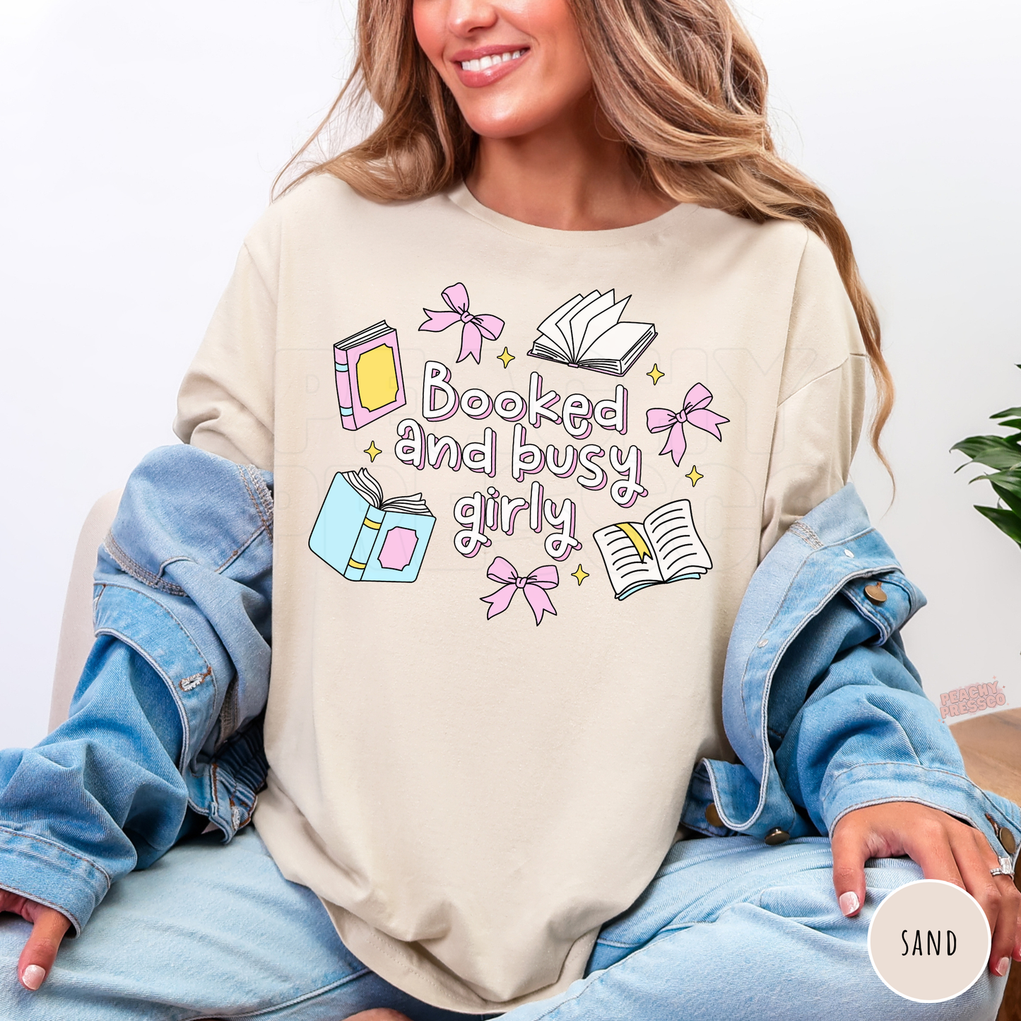 Woman wearing a sand-colored crewneck sweatshirt featuring a cute "Booked and Busy Girly" graphic with pastel books, pink bows, open books, and sparkles. Oversized cozy sweatshirt styled with a light denim jacket and light wash jeans.