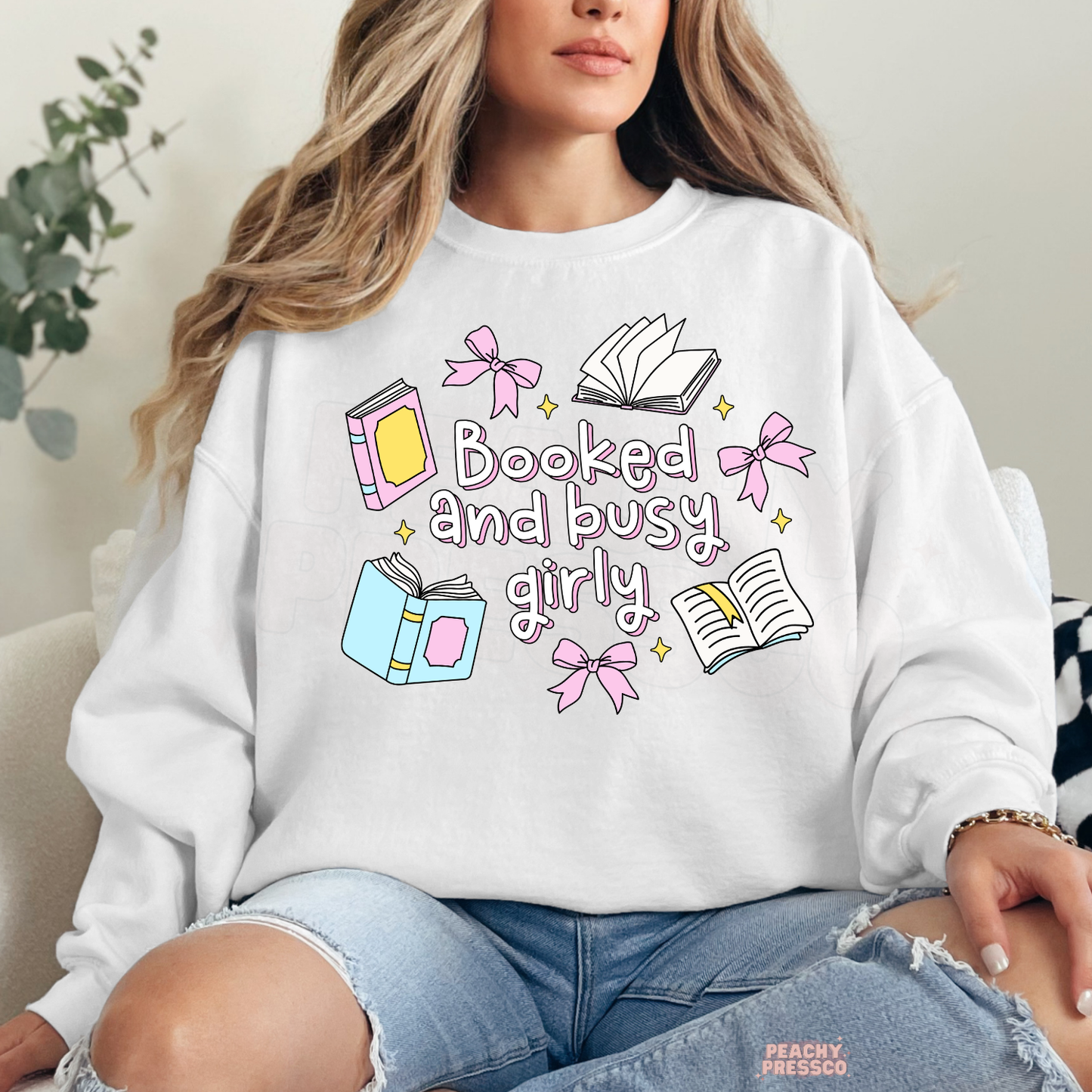White crewneck sweatshirt featuring "Booked and Busy Girly" with pastel books, bows, open books, and cute book lover illustrations.
