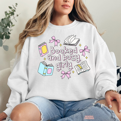 White crewneck sweatshirt featuring "Booked and Busy Girly" with pastel books, bows, open books, and cute book lover illustrations.
