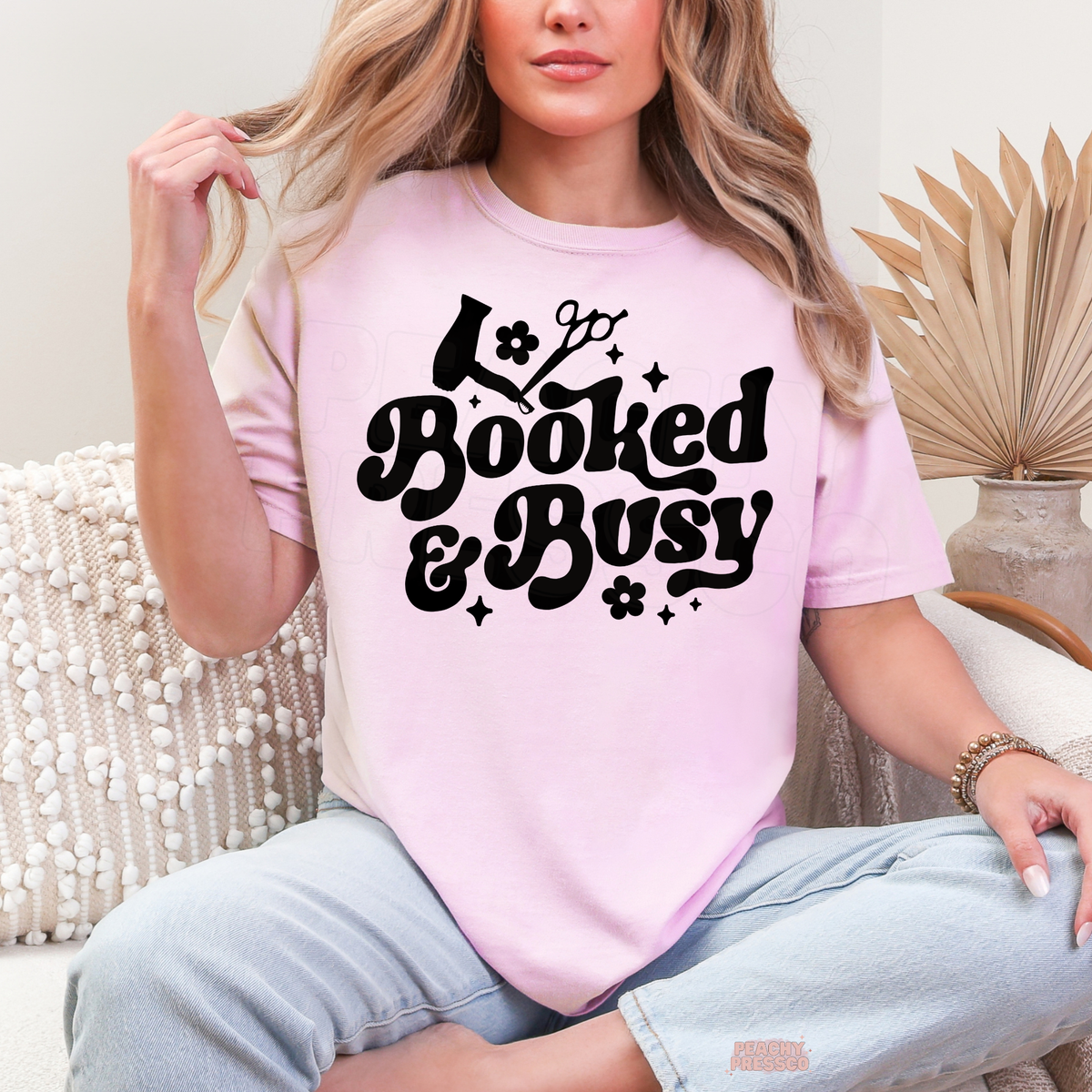 Woman wearing a light pink Booked & Busy hairstylist T-shirt featuring black retro lettering, hair dryer, scissors, flower illustrations and sparkles.