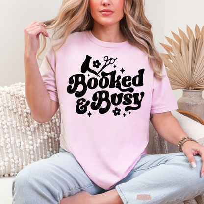 Woman wearing a light pink Booked & Busy hairstylist T-shirt featuring black retro lettering, hair dryer, scissors, flower illustrations and sparkles.