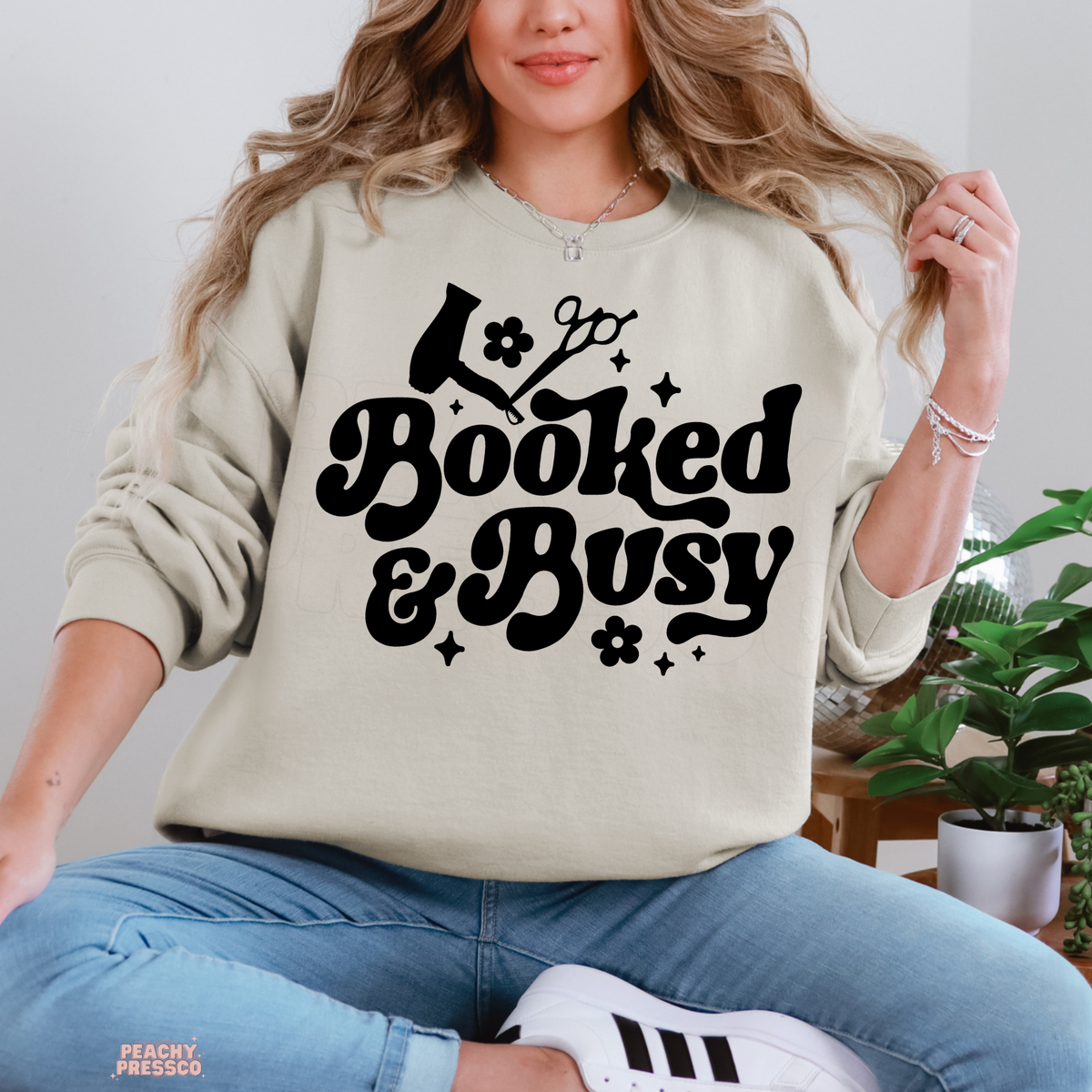 Woman wearing a sand Booked & Busy hairstylist crewneck sweatshirt featuring black vintage lettering, hair dryer, scissors and floral accents.