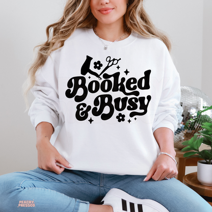 Woman wearing a white Booked & Busy hairstylist sweatshirt with bold retro lettering, black hair dryer and scissors illustrations, flowers and stars.