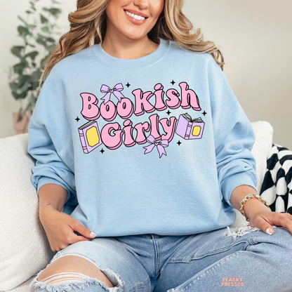 Woman wearing a light blue crewneck sweatshirt featuring a pastel "Bookish Girly" graphic with pink lettering, lavender bows, books, and sparkles. Cozy oversized sweatshirt perfect for book lovers.