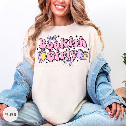 Woman wearing a natural-colored crewneck sweatshirt featuring a pink "Bookish Girly" graphic with pastel books, lavender bows, and sparkles. Cozy oversized sweatshirt styled with a light denim jacket.