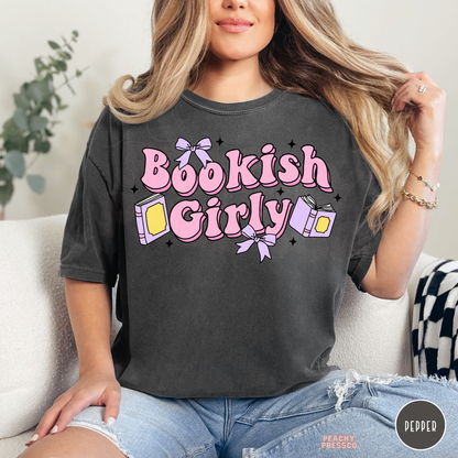 Woman wearing a pepper Comfort Colors t-shirt featuring a pink "Bookish Girly" graphic with lavender bows, pastel books, and sparkles. Vintage-inspired oversized tee for readers.