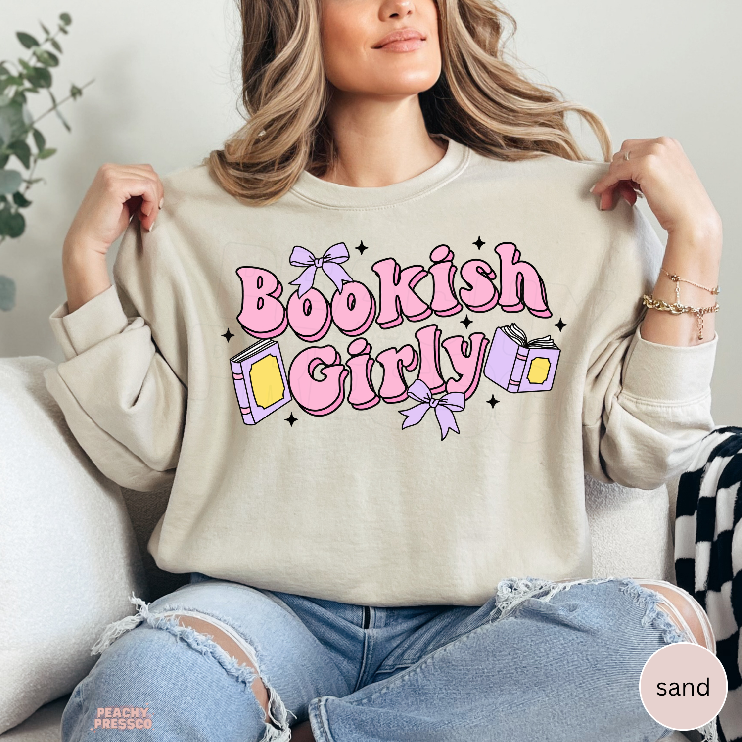 Woman wearing a sand-colored crewneck sweatshirt featuring a whimsical "Bookish Girly" graphic with pink lettering, books, lavender bows, and sparkles. Oversized cozy sweatshirt for book lovers.