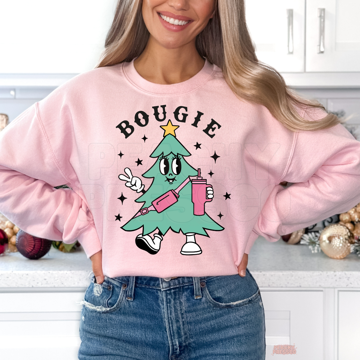 Woman standing in a kitchen wearing a light pink crewneck sweatshirt featuring a retro cartoon Bougie Christmas tree holding a tumbler cup.