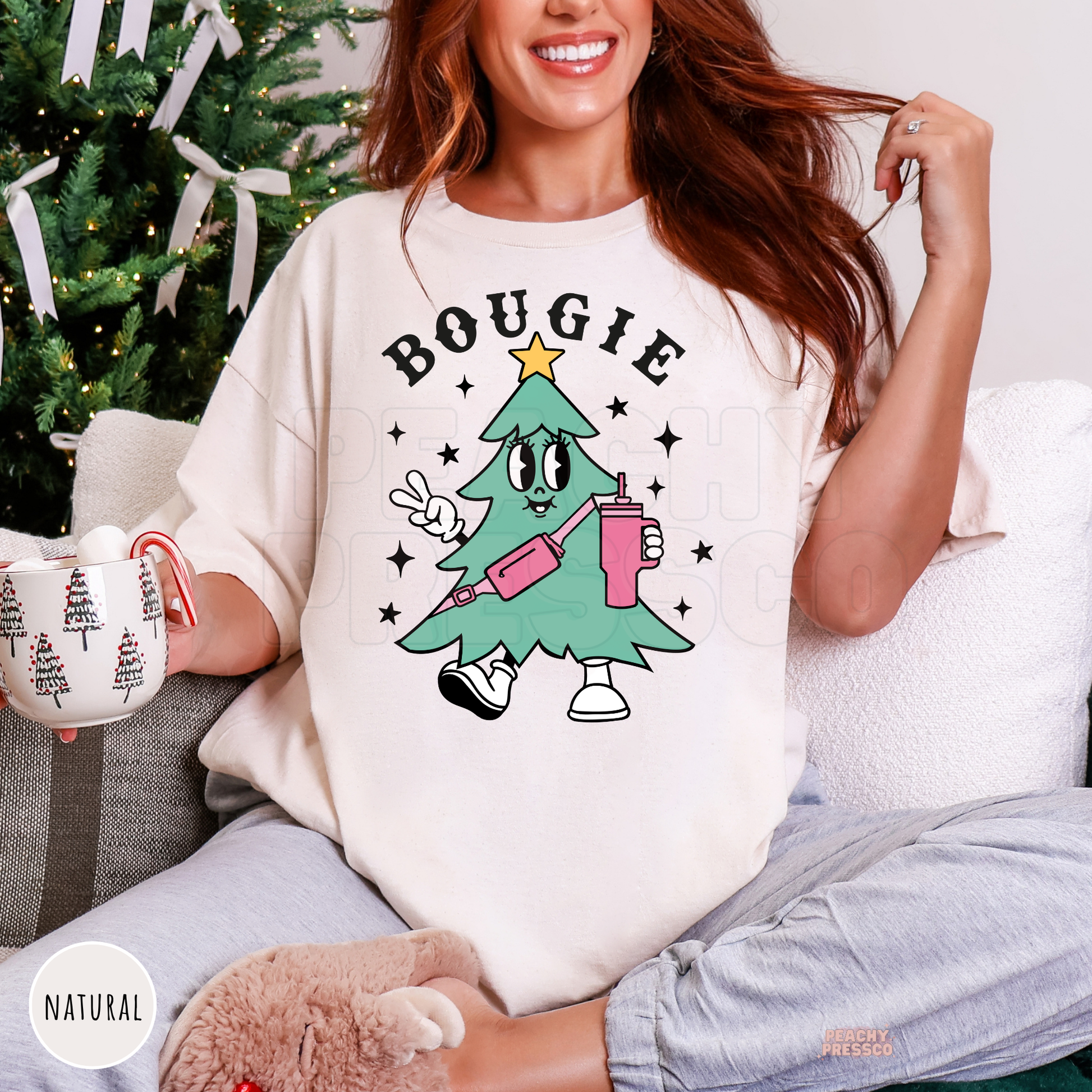 Woman holding a holiday mug wearing a natural cream oversized shirt featuring a retro Bougie Christmas tree wearing a pink belt bag.
