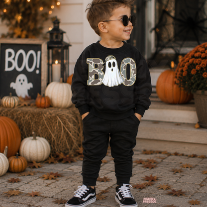 Boy wearing a black Boo camo ghost Halloween sweatshirt with sunglasses surrounded by pumpkins and fall decor.