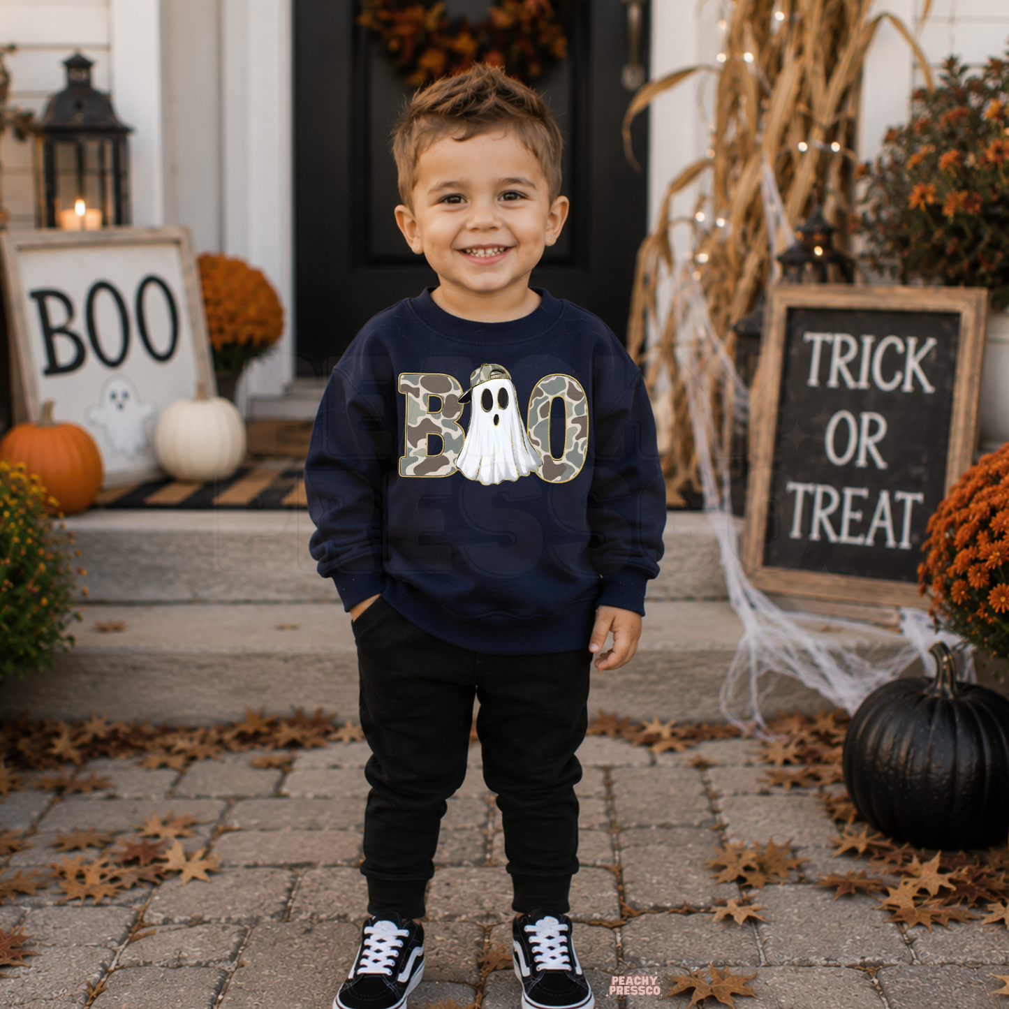 Boy wearing a navy Boo camo ghost Halloween sweatshirt with black pants in a festive pumpkin porch setting.