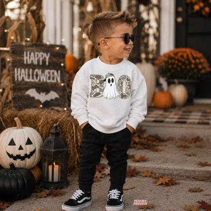 Boy wearing a white Boo camo ghost Halloween sweatshirt with black pants in a festive outdoor Halloween setting.