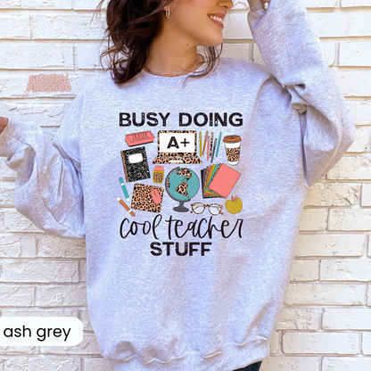 Ash grey teacher sweatshirt featuring a "Busy Doing Cool Teacher Stuff" graphic with classroom essentials, leopard print accessories, coffee cup, globe, notebooks, pencils, and apple.