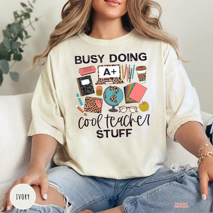 Ivory Comfort Colors teacher t-shirt with a "Busy Doing Cool Teacher Stuff" graphic featuring classroom supplies, leopard print details, globe, coffee cup, and school accessories.