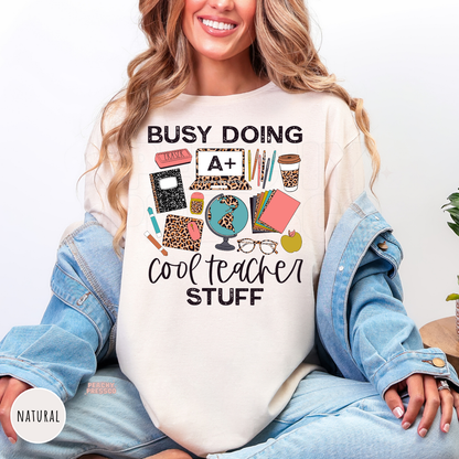 Natural teacher shirt featuring "Busy Doing Cool Teacher Stuff" with classroom essentials including notebooks, globe, coffee cup, pencils, laptop, apple, glasses, and leopard print accents.