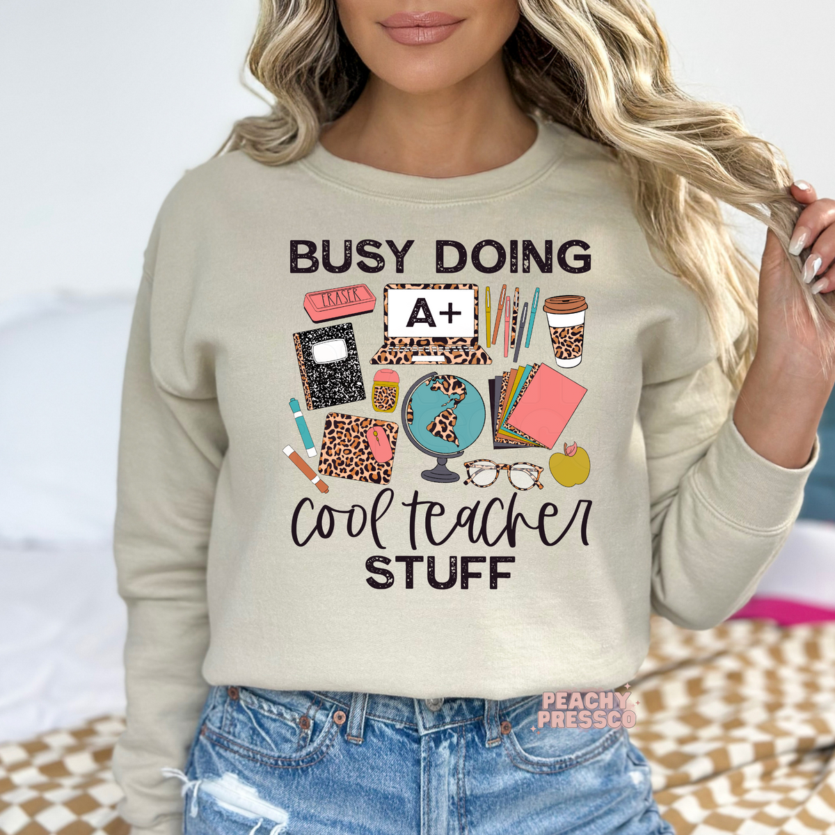 Sand teacher sweatshirt with a "Busy Doing Cool Teacher Stuff" design featuring colorful classroom supplies, leopard print notebook and coffee cup, globe, laptop, and apple.