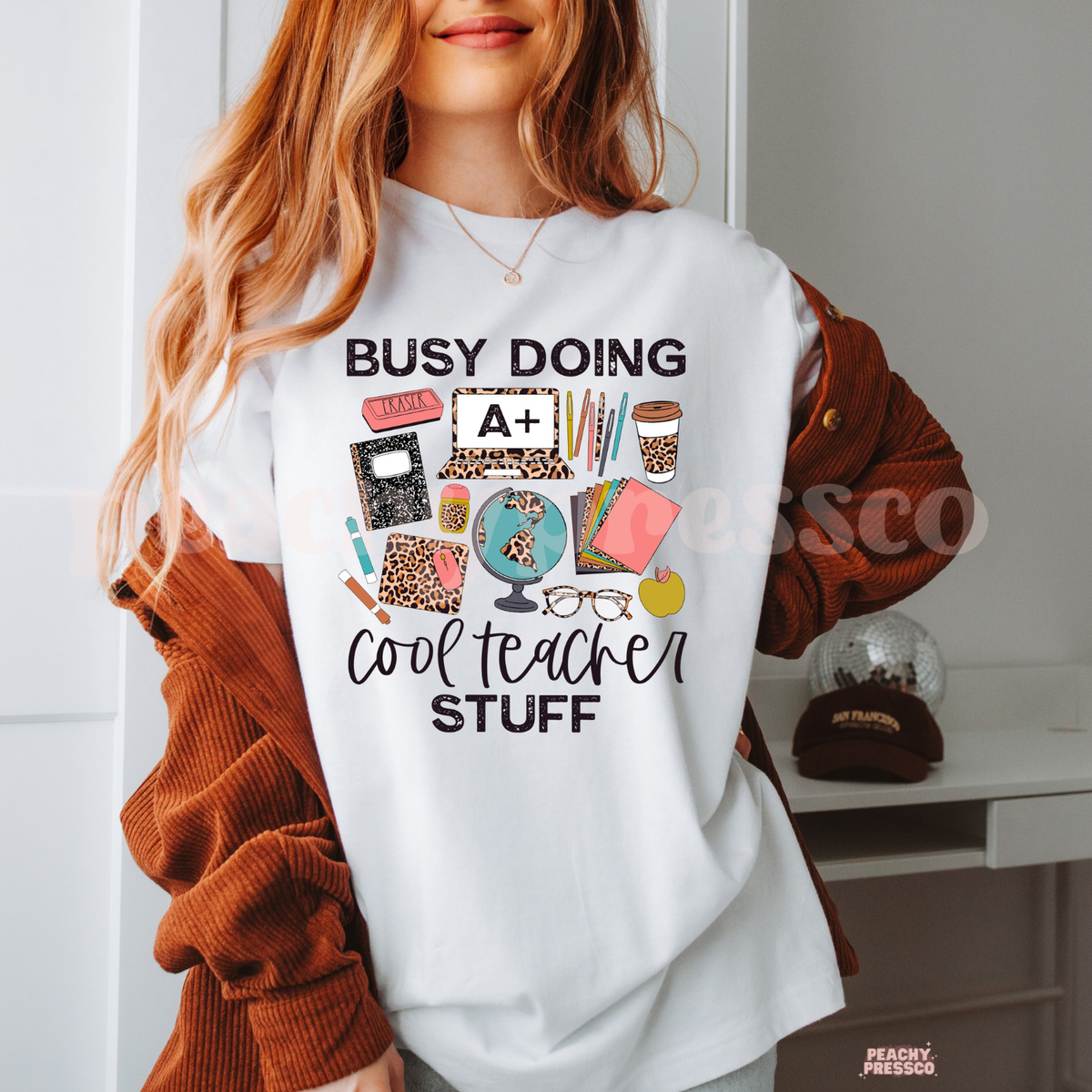 White teacher t-shirt featuring "Busy Doing Cool Teacher Stuff" with school supplies, globe, laptop, coffee cup, pens, notebooks, glasses, and leopard print accents.