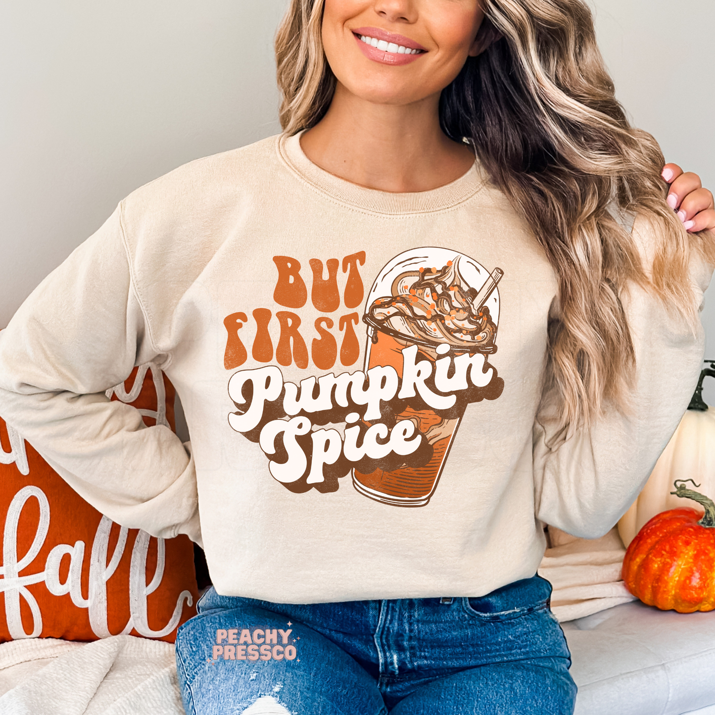 Woman wearing a sand crewneck sweatshirt featuring a vintage But First Pumpkin Spice iced coffee graphic in warm orange and brown fall tones.