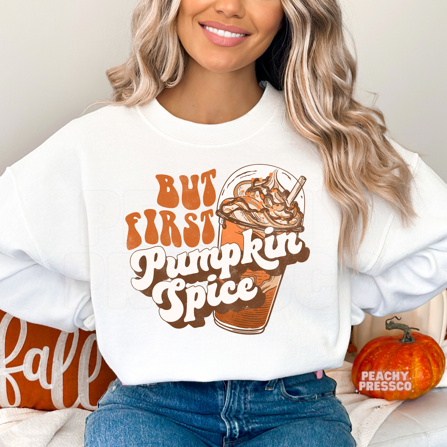 Woman wearing a white crewneck sweatshirt featuring a retro "But First Pumpkin Spice" graphic with an iced pumpkin spice coffee in vintage fall colors.