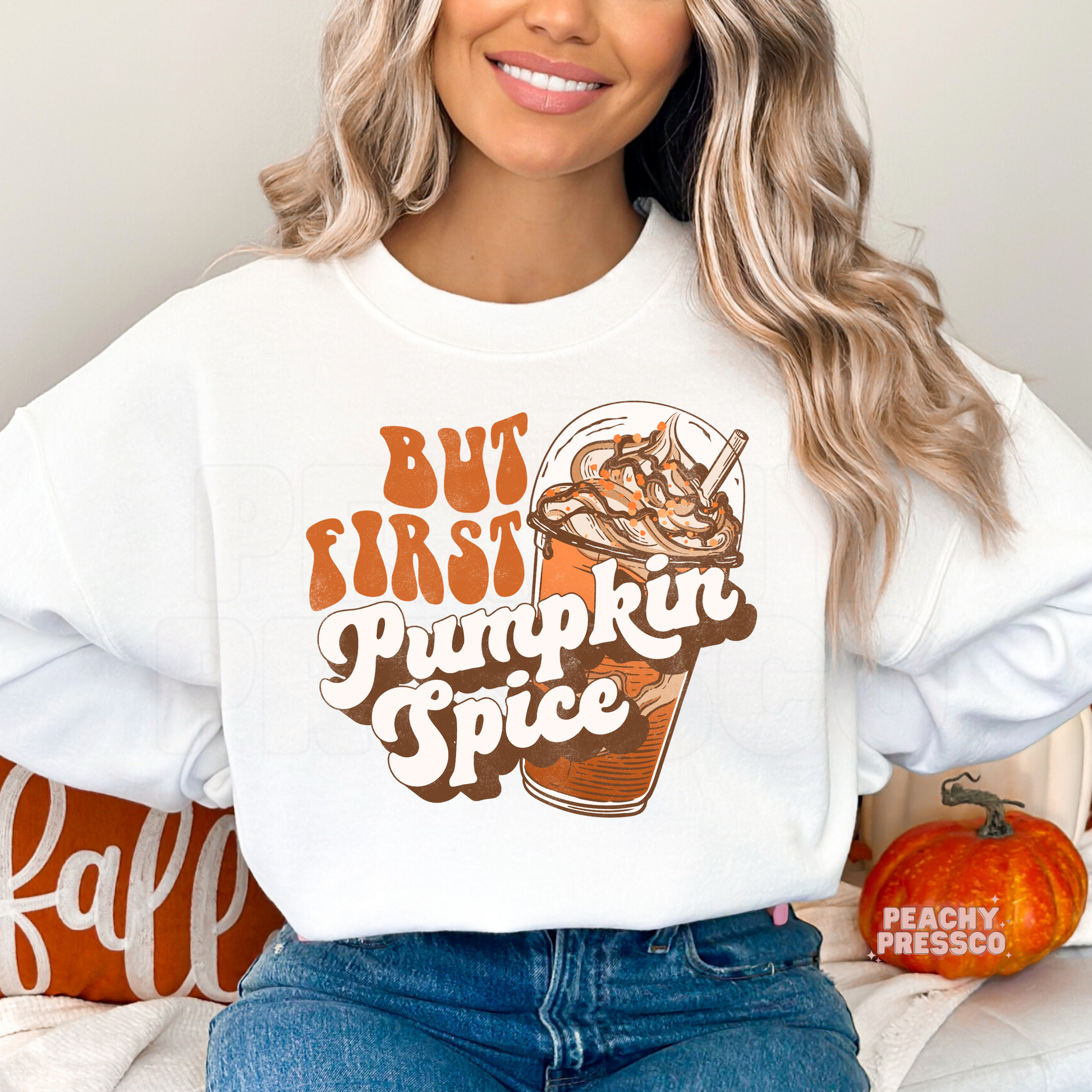 Woman wearing a white crewneck sweatshirt featuring a retro "But First Pumpkin Spice" graphic with an iced pumpkin spice coffee in vintage fall colors.