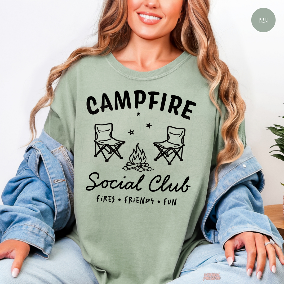 Bay green Campfire Social Club shirt with a minimalist camping illustration, bonfire, folding chairs and outdoor adventure quote.