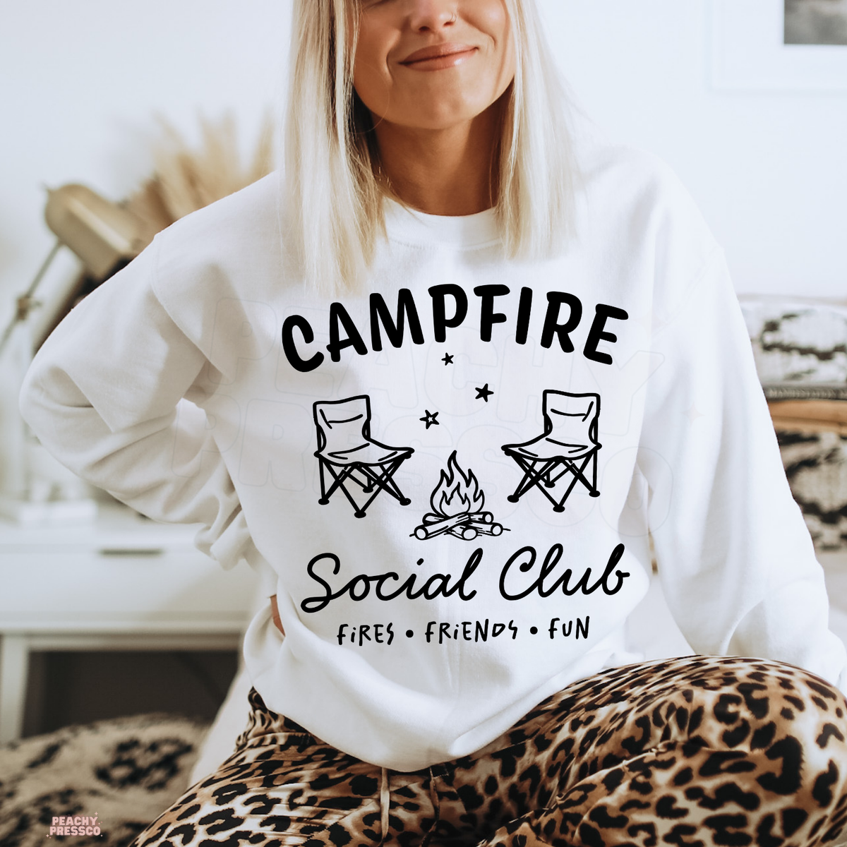 White Campfire Social Club sweatshirt with a campfire illustration, folding camping chairs and playful outdoor adventure lettering.
