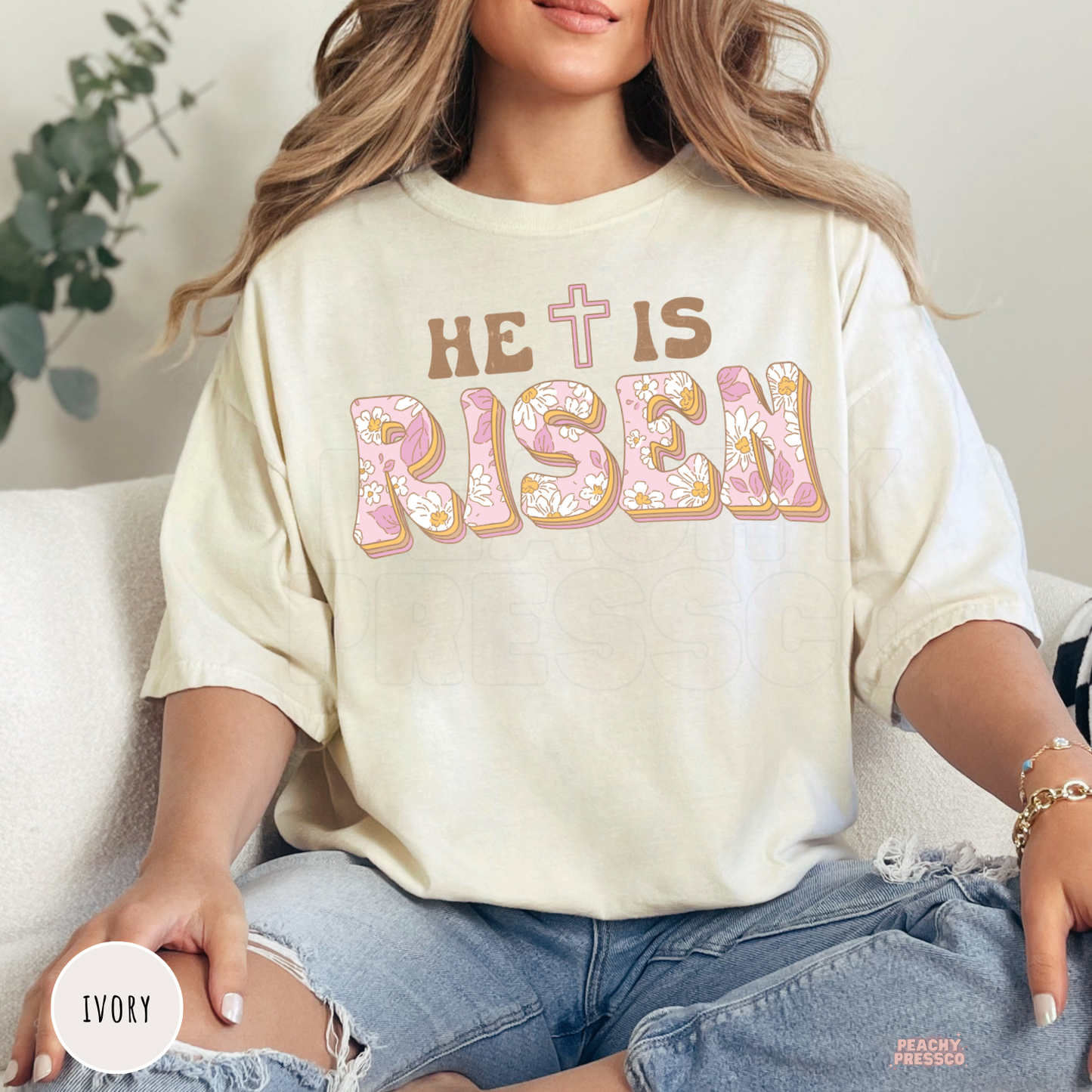 Ivory unisex t-shirt featuring a retro floral "He Is Risen" graphic with pastel pink flowers, brown lettering, and a cross. Soft Christian Easter tee celebrating the resurrection of Jesus.
