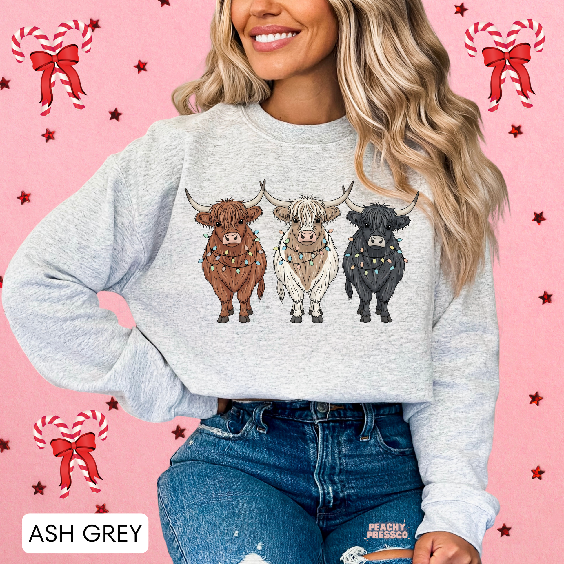 A woman wearing an ash grey crewneck sweatshirt with a graphic of three Highland cows decorated with Christmas lights, set against a pink holiday-themed background with candy canes and stars.