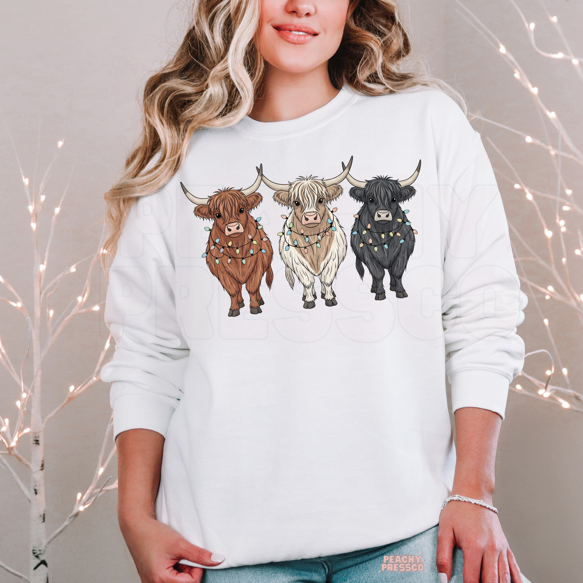 A woman wearing a white crewneck sweatshirt with a graphic of three Highland cows in brown, white, and black, posed in front of a soft-focus background with warm string lights.