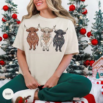 Woman wearing an ivory short sleeve t-shirt with three festive Highland cows wrapped in Christmas lights, sitting in front of Christmas trees.