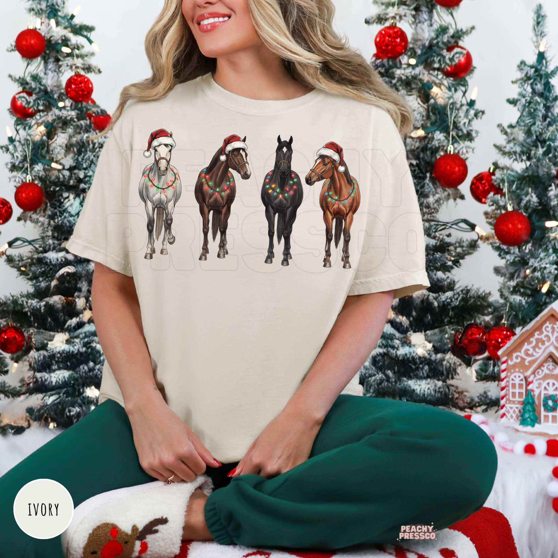 A woman wearing a ivory colored shirt with a graphic of four horses in Santa hats, posed in front of decorated Christmas trees.