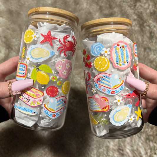 Coastal-themed 16oz glass cup with bamboo lid featuring colorful sardines, lemons, shells, crabs, and Mediterranean-inspired illustrations held in hand.