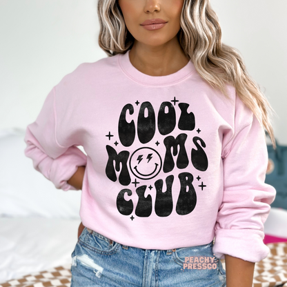 Light pink sweatshirt featuring the "Cool Moms Club" retro graphic with a smiley face and bold vintage lettering. Fun gift for moms.
