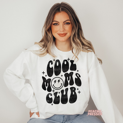 White sweatshirt featuring a retro "Cool Moms Club" graphic with a smiley face and vintage black lettering. Trendy graphic sweatshirt for stylish moms.
