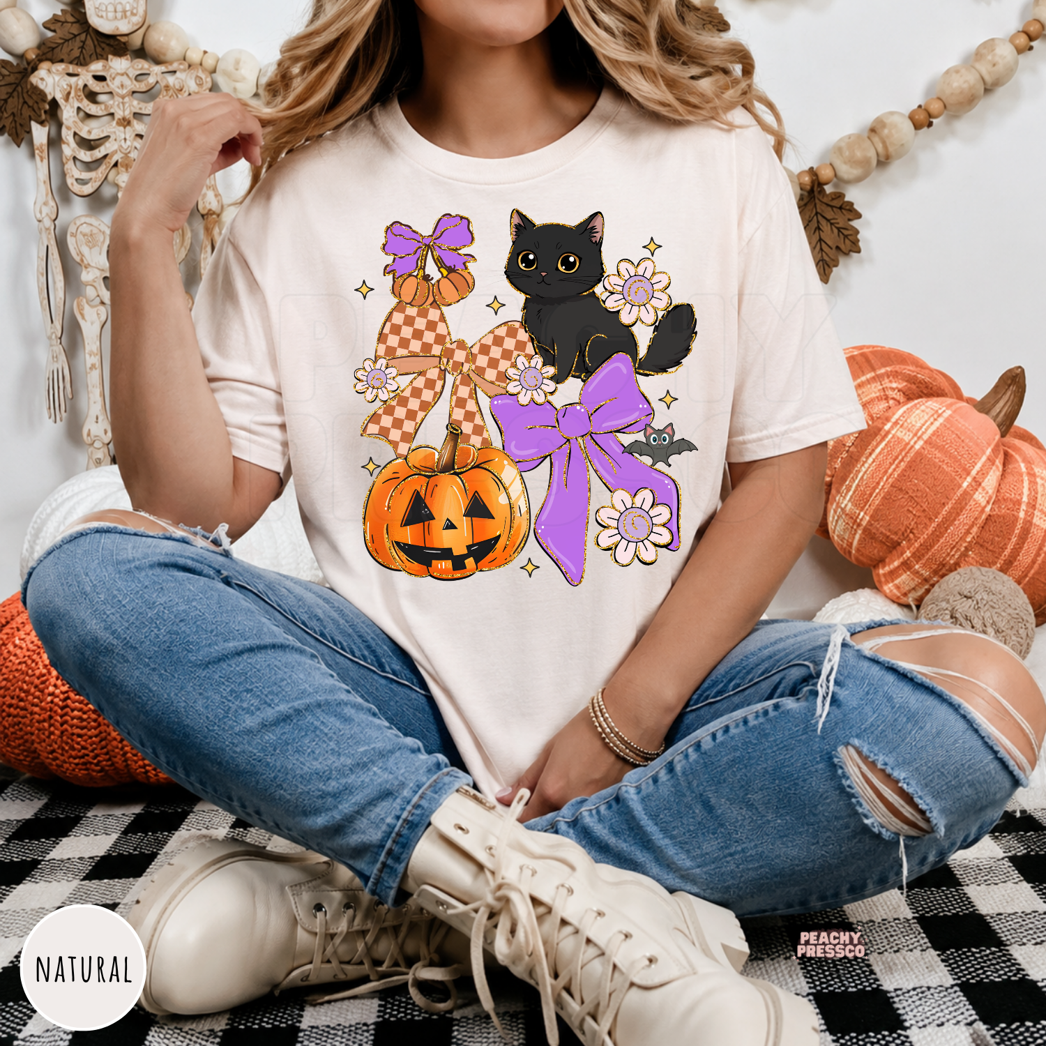 Woman wearing a natural Halloween graphic tee with a black cat, jack-o'-lantern pumpkin, purple bows, flowers and bat design.