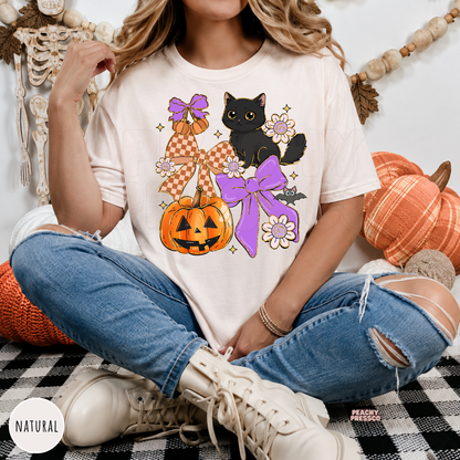 Woman wearing a natural Halloween graphic tee with a black cat, jack-o'-lantern pumpkin, purple bows, flowers and bat design.