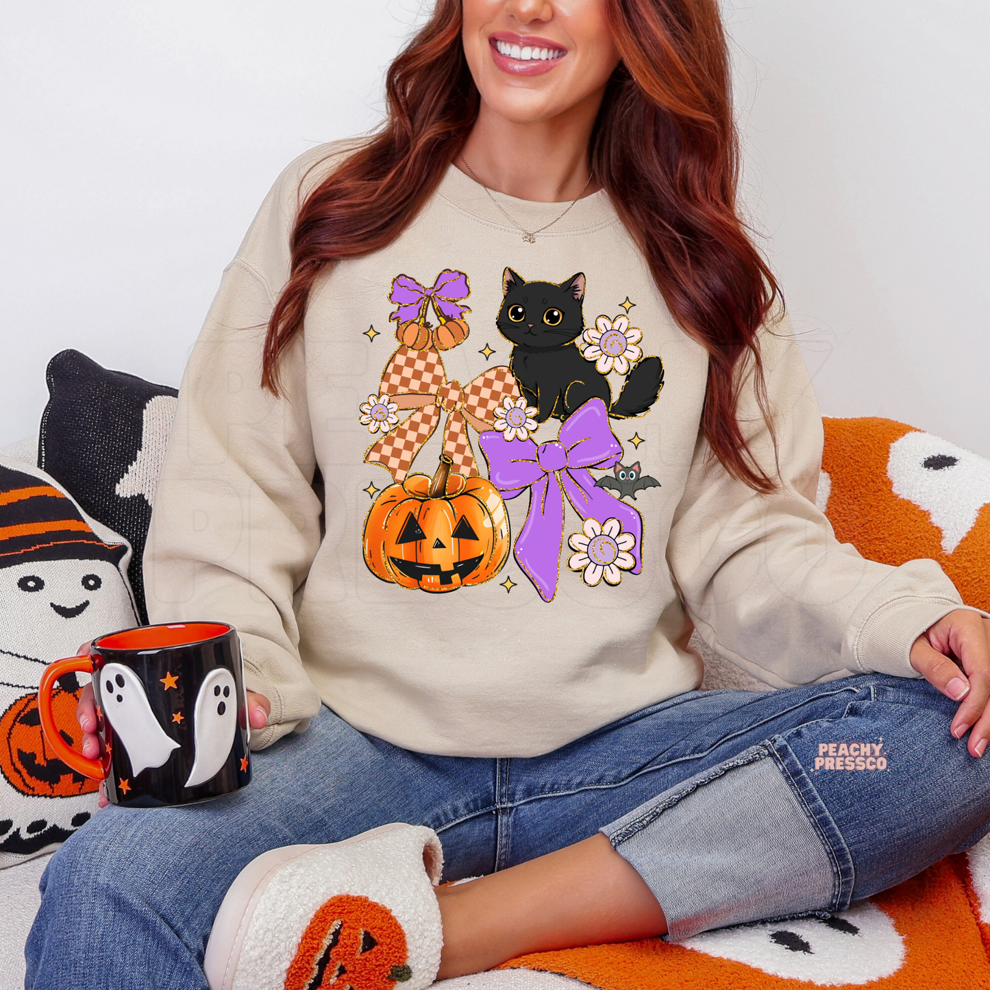 Woman wearing a sand sweatshirt with a cute Halloween black cat, pumpkin, purple coquette bows, flowers and bat design.
