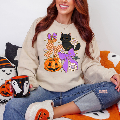 Woman wearing a sand sweatshirt with a cute Halloween black cat, pumpkin, purple coquette bows, flowers and bat design.