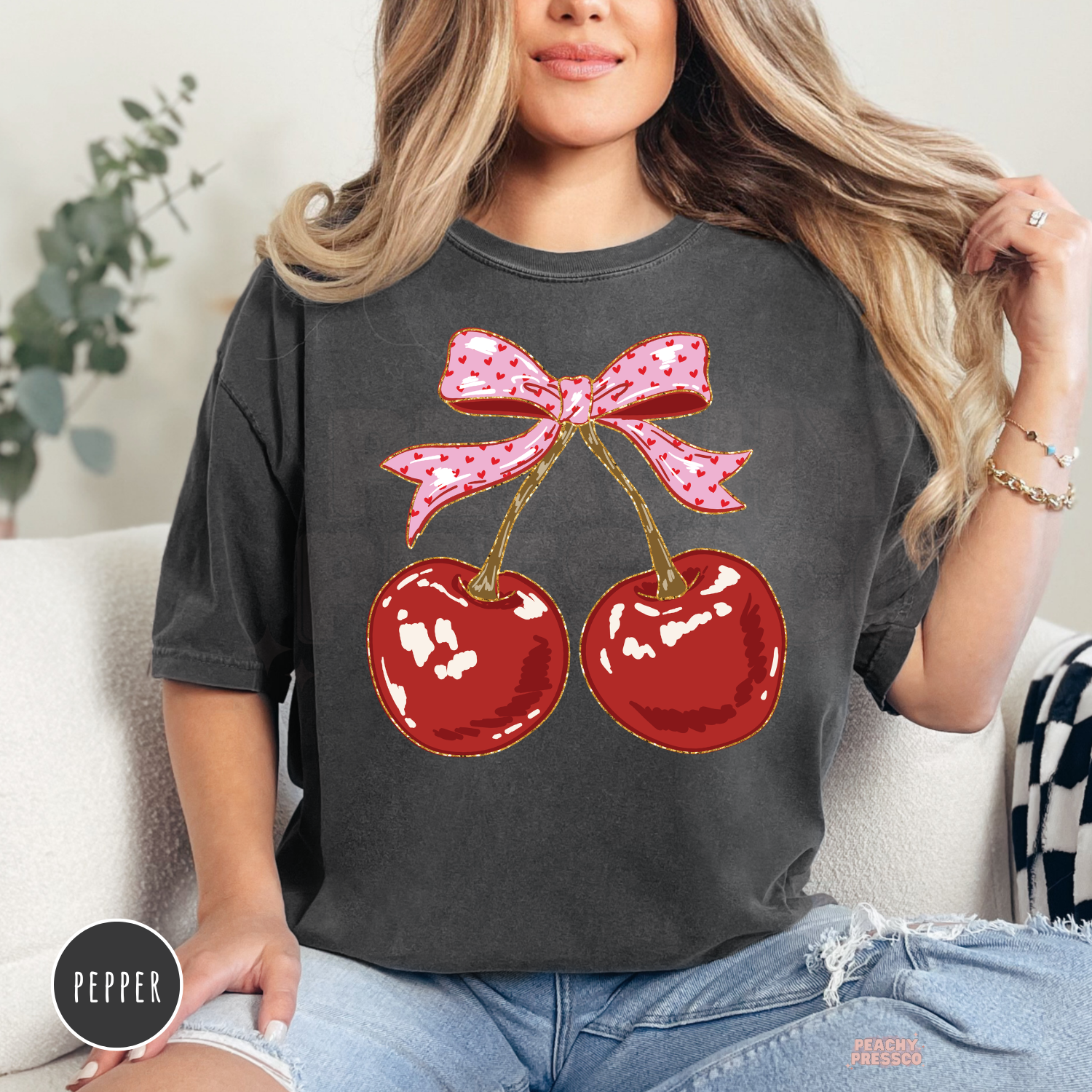 Woman wearing a pepper Comfort Colors graphic t-shirt featuring two red cherries with a pink coquette bow, cute aesthetic cherry shirt.