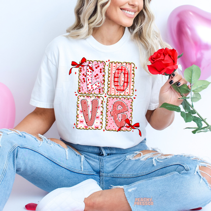White graphic tee with a colorful LOVE patchwork design featuring heart prints, gingham patterns, red bows, and playful Valentine's details.