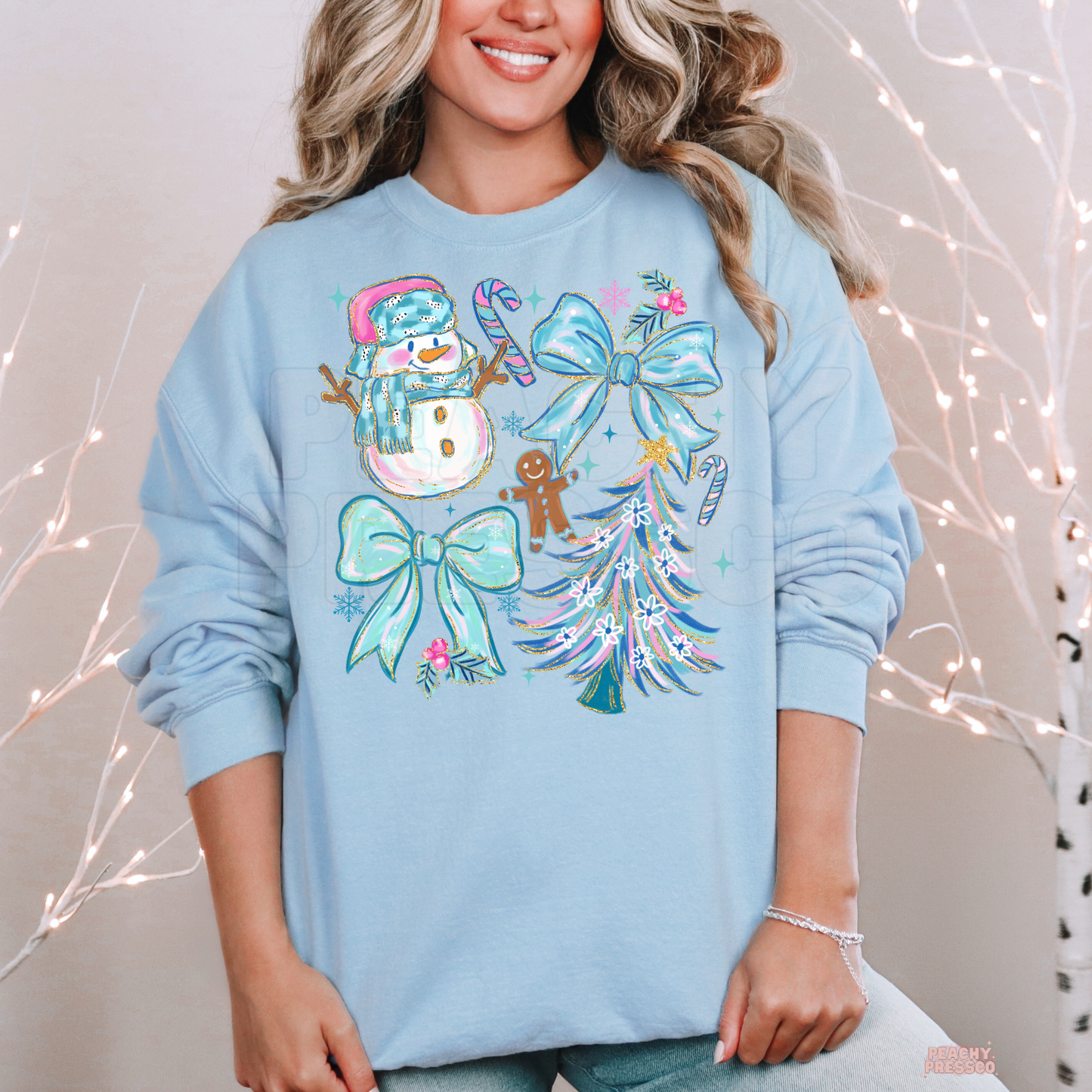 A woman wearing a light blue crewneck sweatshirt with a colorful Christmas graphic print featuring a snowman, candy canes, bows, a gingerbread man, and a decorated tree, set against a warm neutral background with decorative string lights.