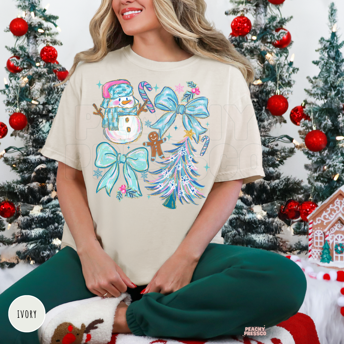 A woman wearing an ivory holiday graphic t-shirt with a snowman, candy canes, bows, and a Christmas tree design, paired with dark green pants, sitting in front of decorated Christmas trees.