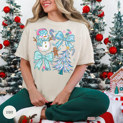 A woman wearing an ivory holiday graphic t-shirt with a snowman, candy canes, bows, and a Christmas tree design, paired with dark green pants, sitting in front of decorated Christmas trees.