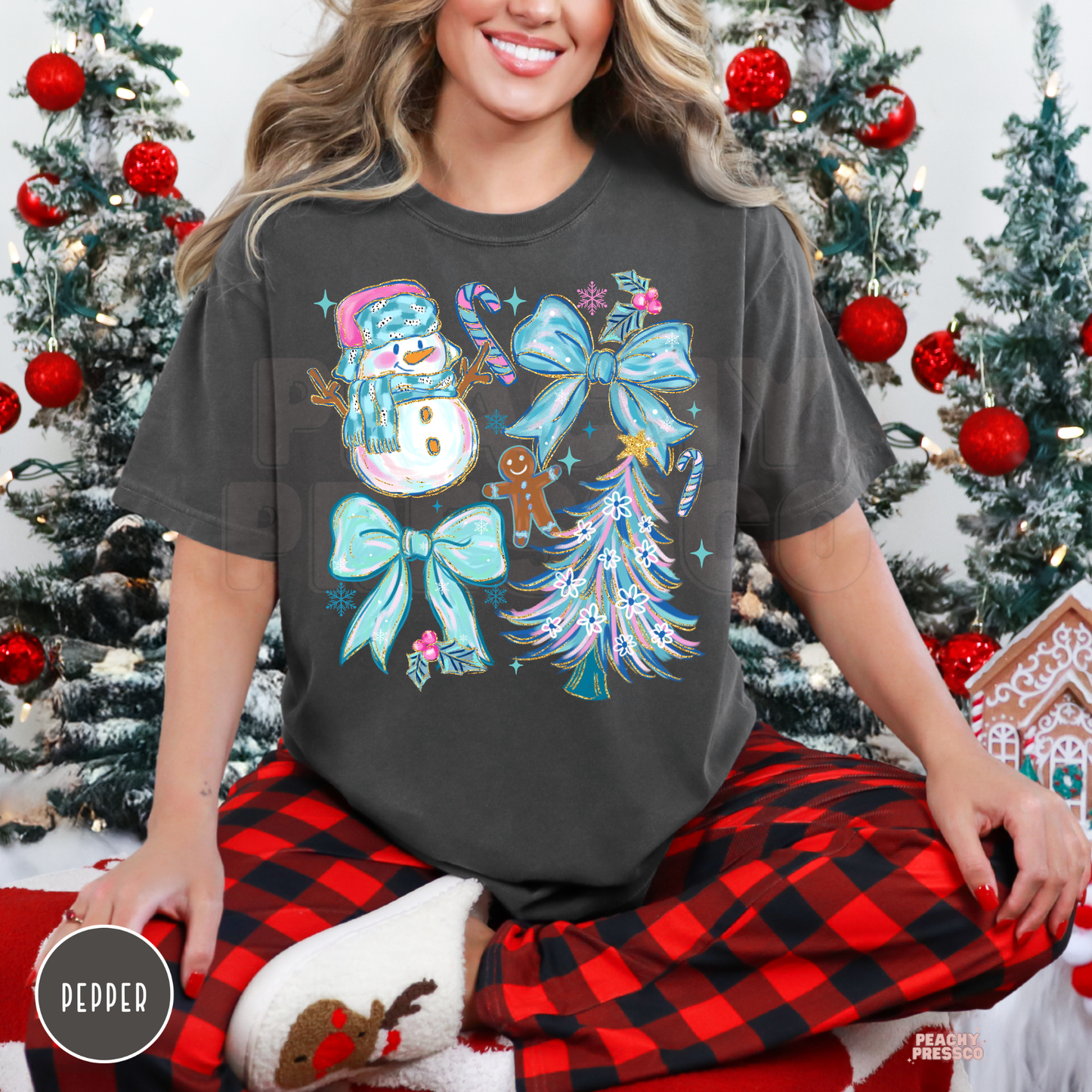 A woman wearing comfort colors shirt in Pepper with a colorful Christmas graphic print featuring a snowman, candy canes, gingerbread man, bows, and a decorated tree. She is sitting in front of a decorated Christmas tree, wearing red and black buffalo plaid pajama pants and white slippers.